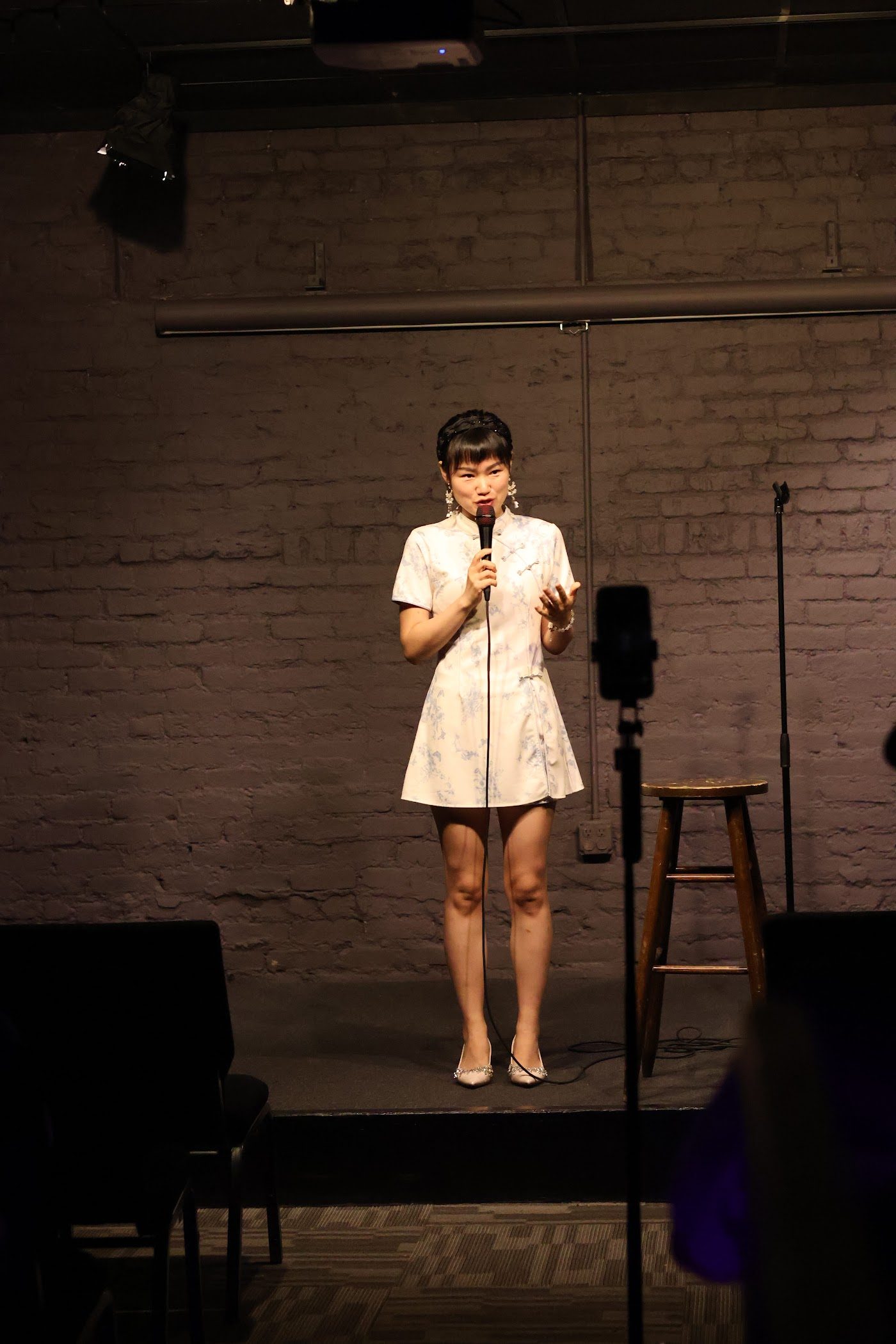 Ginger Phoenix performing stand-up on a small stage with a brick wall backdrop